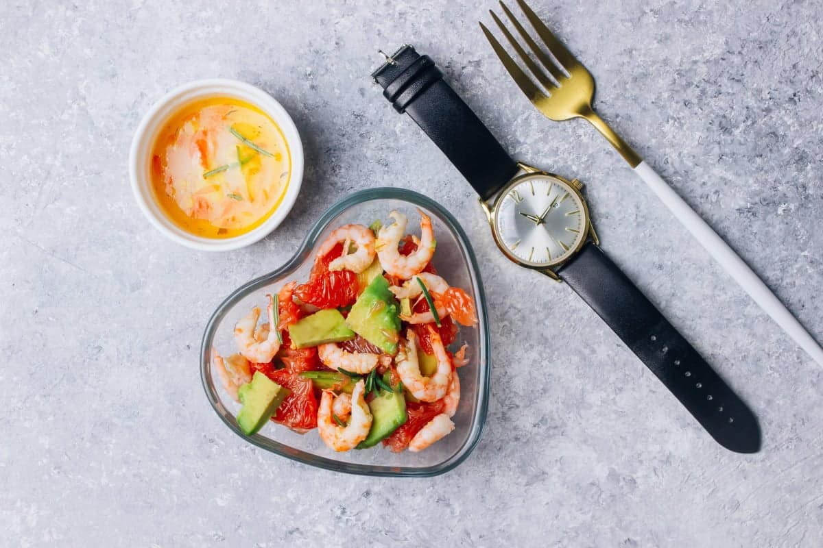 a watch to time Intermittent Fasting with a fork, and shrimp avocado salad in a heart shaped bowl