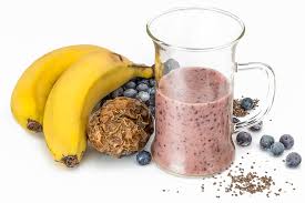 protein shake in a glass pitcher with bananas, blueberries, and granola next to it