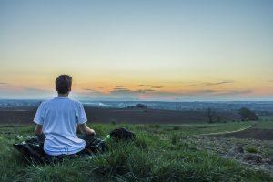 A man sitting on grass facing the skyline and meditating to improve focus