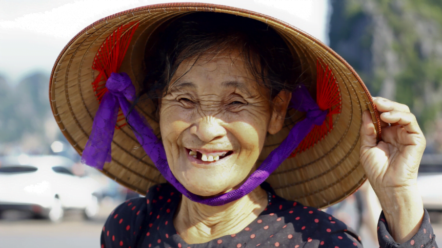An asian woman that is missing teeth is wearing a straw hat and smiling