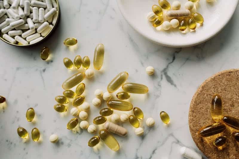 A variety of supplement pills scattered on a white marble table, including capsules, tablets, and softgels.