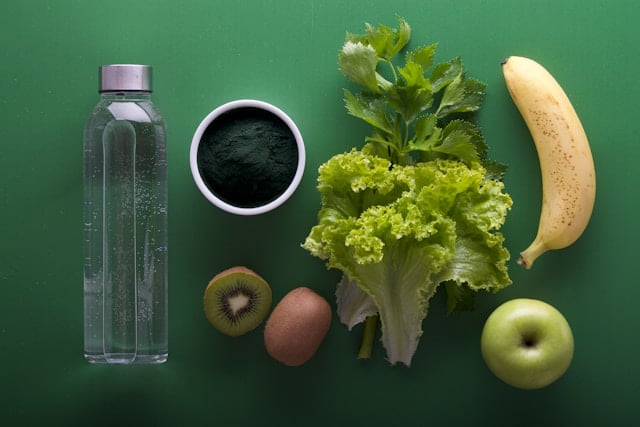 Flat lay of a water bottle, banana, kiwi, spirulina, and leafy greens—nutrient-rich foods for concussion recovery. These foods support brain healing with hydration, antioxidants, and anti-inflammatory properties. Learn more at Nutrition Therapy Institute.