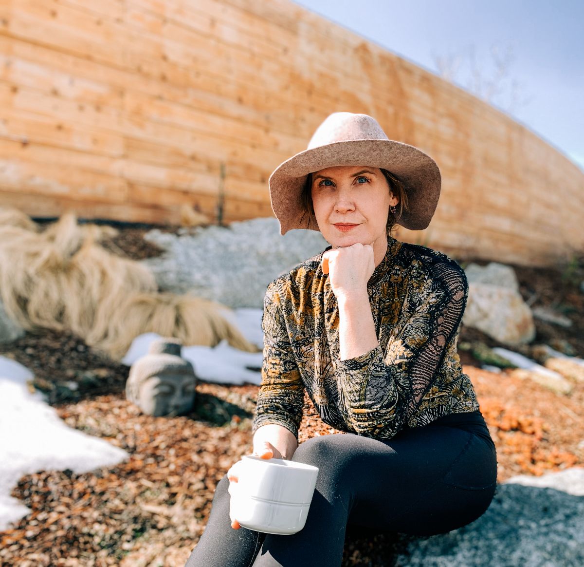 Carla Abate sitting in front of a rock formation, wearing a hat, and holding a mug