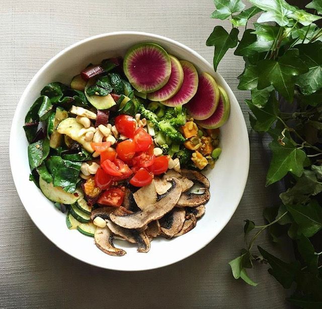 A green salad with watermelon radish, mushrooms, and bell pepper in a white bowl next to a plant